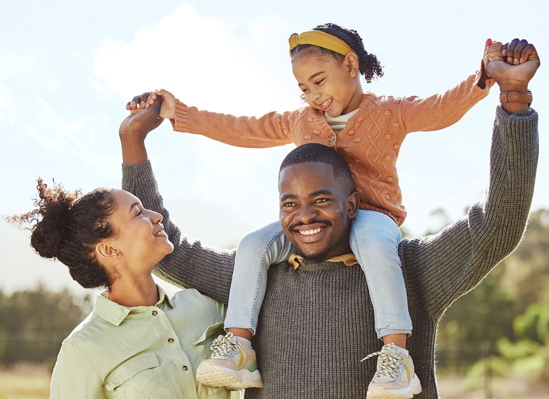 Personal Insurance - Cheerful Father Holding his Young Daughter on his Shoulders While Standing Next to the Mother Outside in the Park